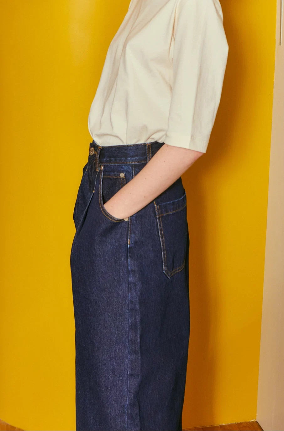 A person wearing a cream short-sleeve blouse and a dark blue denim skirt, standing against a bright yellow wall.