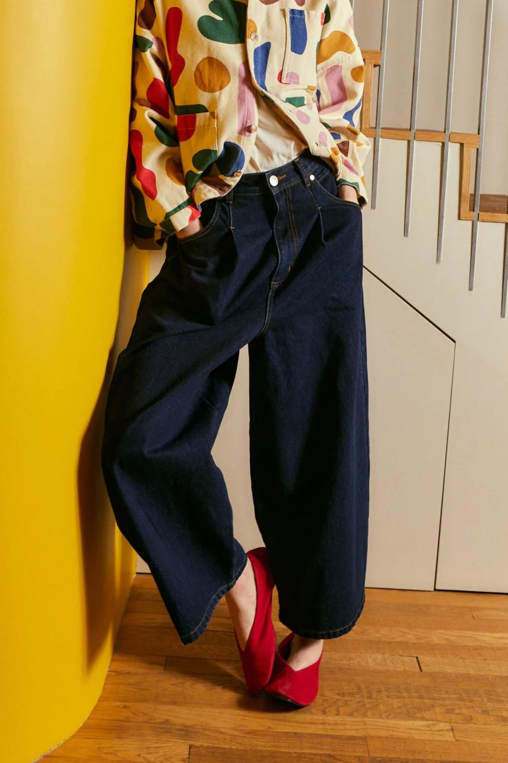A person wearing dark wide-leg denim jeans, a colorful patterned jacket, and red shoes  standing on a wooden floor next to a bright yellow cylindrical wall element.