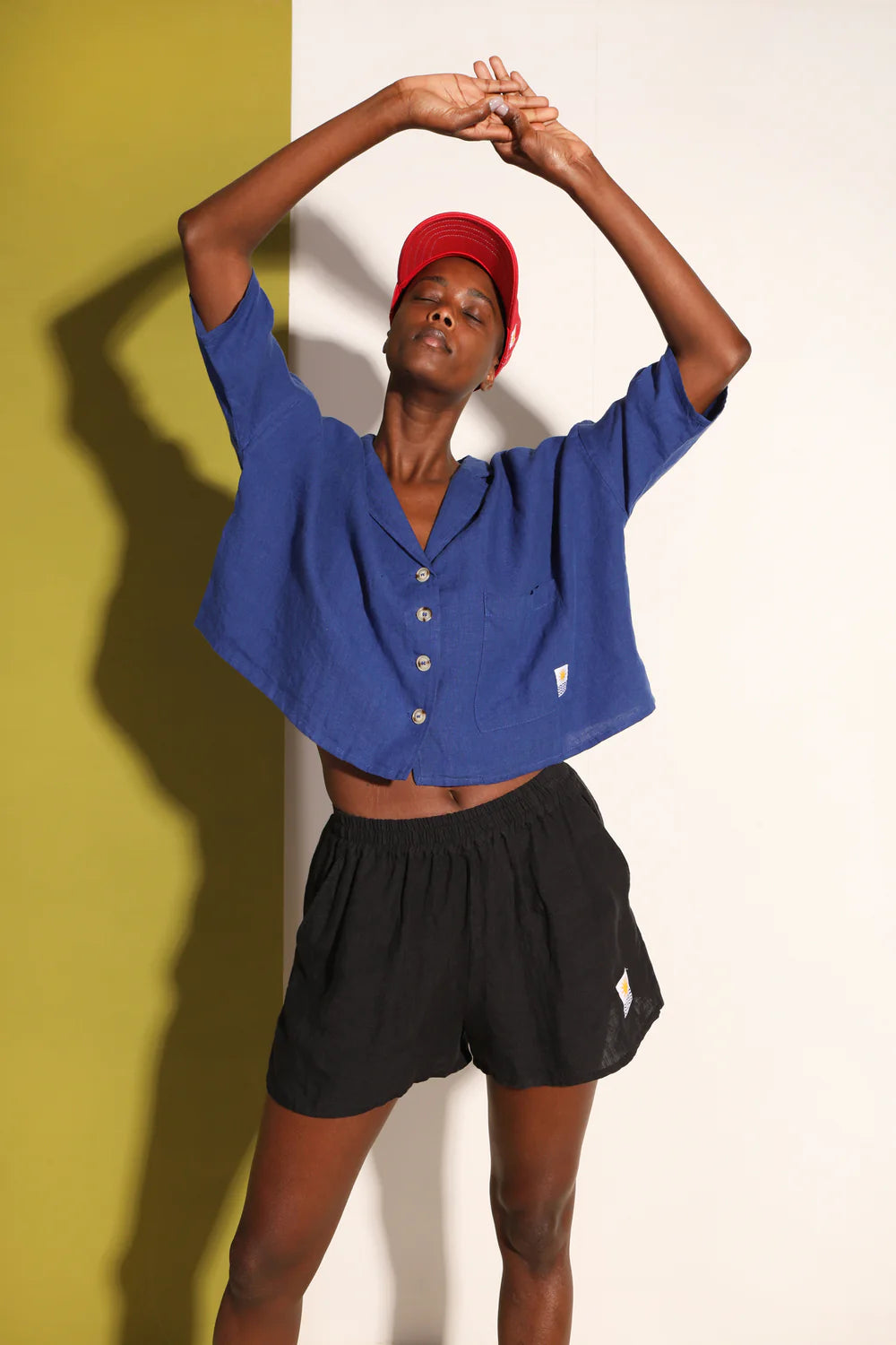 Person wearing a blue shirt and black shorts with a red cap, standing against a yellow and white background.