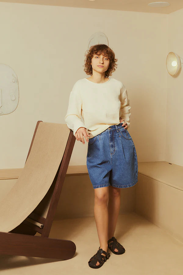 Person wearing a beige sweater and blue denim shorts standing in a minimalistic room.