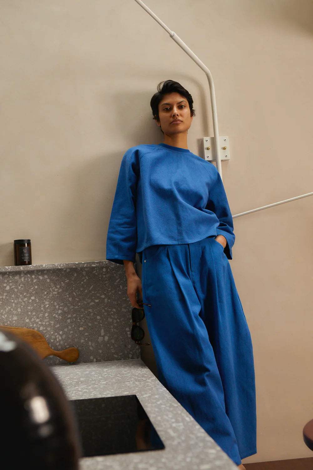 Person wearing a blue LFMarkey top and trousers standing in a room with a textured wall and a shelf.