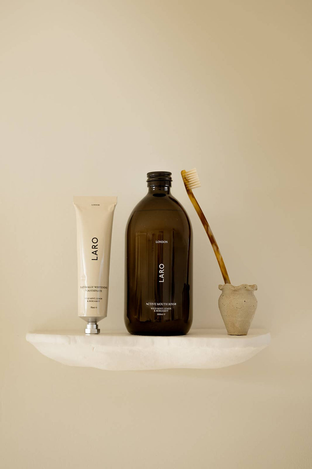 Set of dental products including a brown bottle Laro mouth rinse  with a tube of Laro toothpaste, and brush on a beige surface with a neutral background