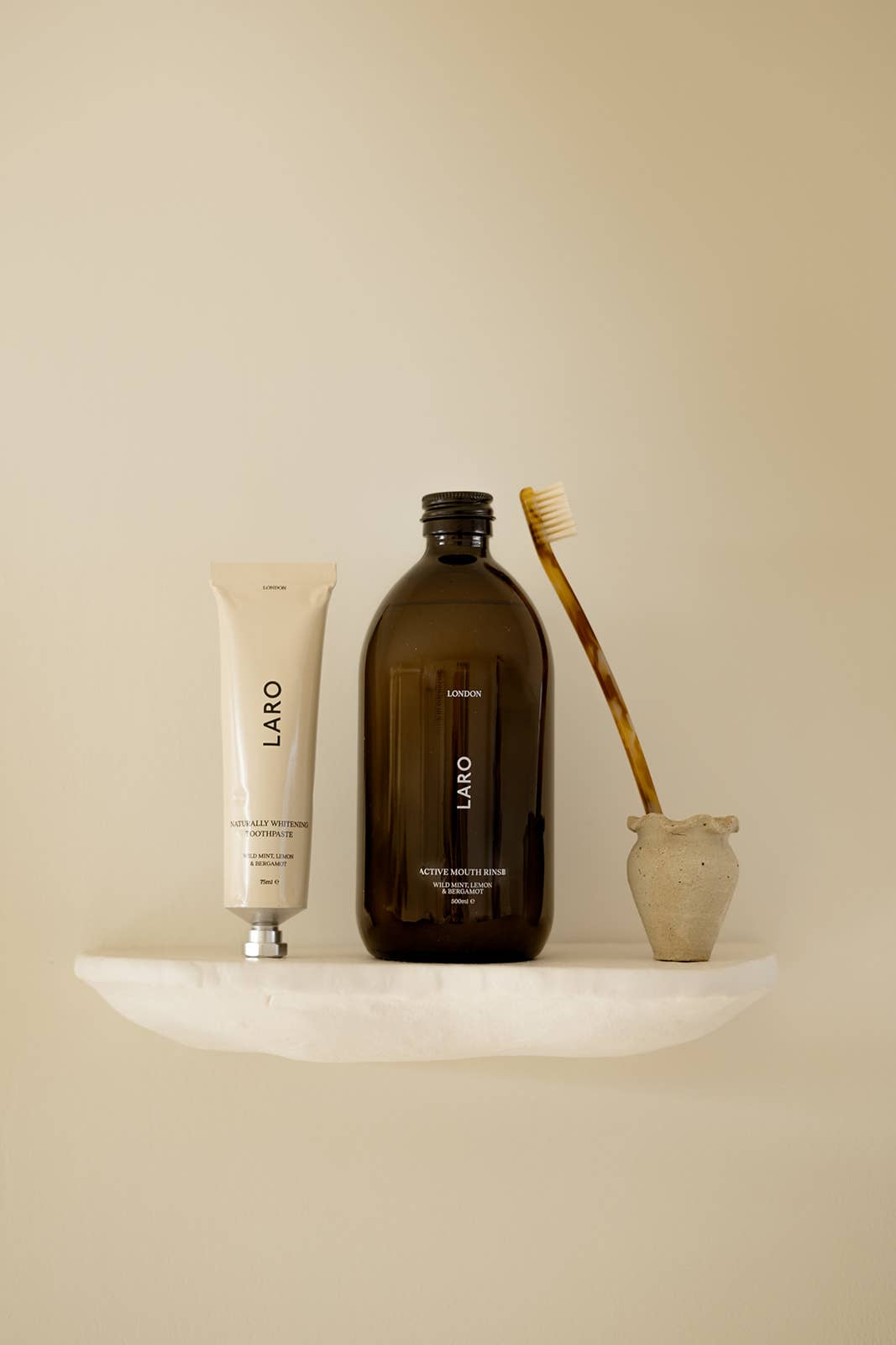 Set of dental products including a brown bottle Laro mouth rinse  with a tube of Laro toothpaste, and brush on a beige surface with a neutral background