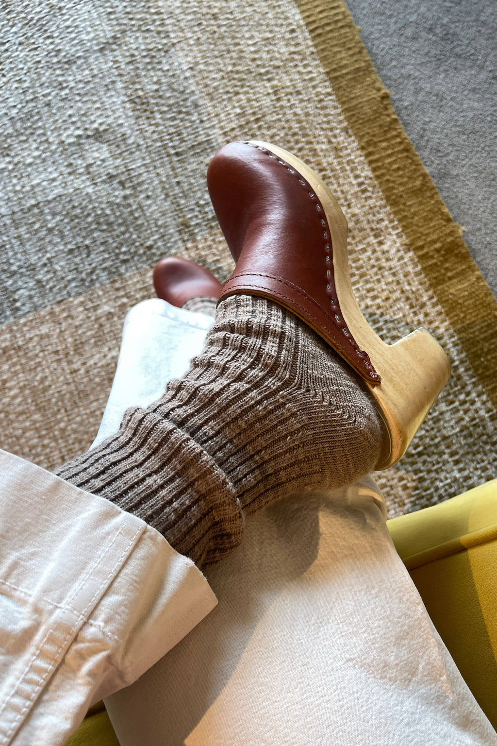 Brown leather clogs with beige soles worn with Dark beige Le Bon Shoppe socks on a textured floor.