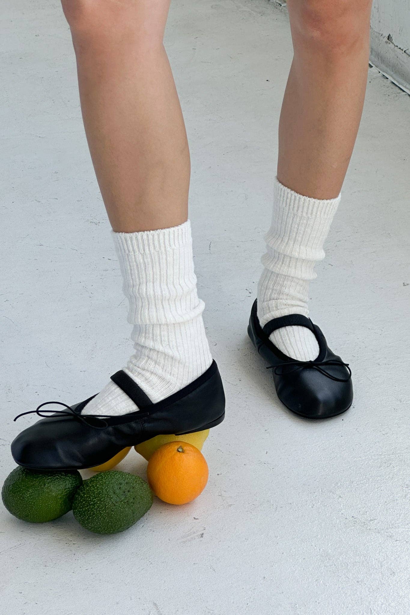 Person wearing black shoes and white Le Bon Shoppe socks with fruits on a light grey floor.