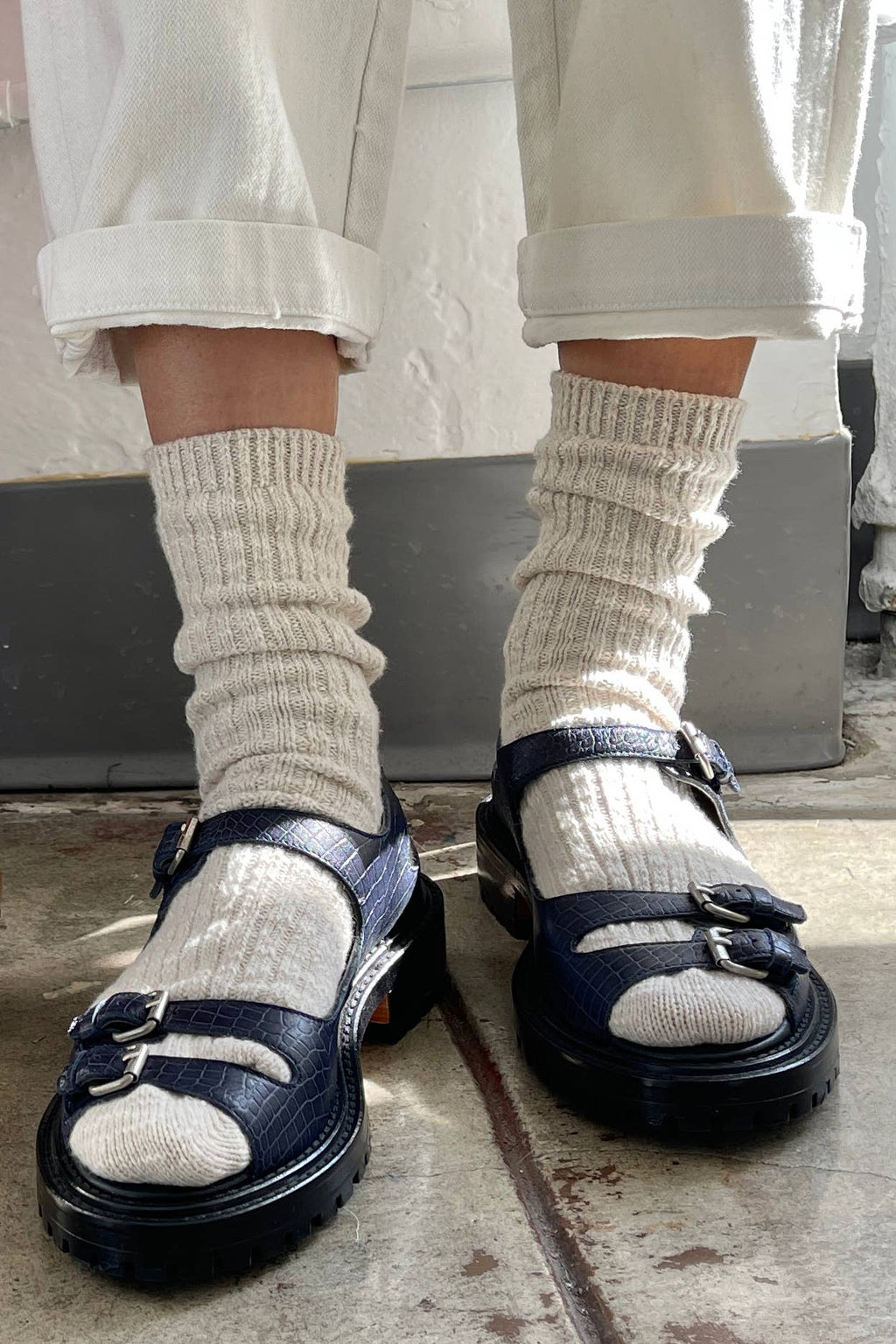 Person wearing navy sandals with beige Le Bon Shoppe  socks and rolled-up pants on a concrete floor.