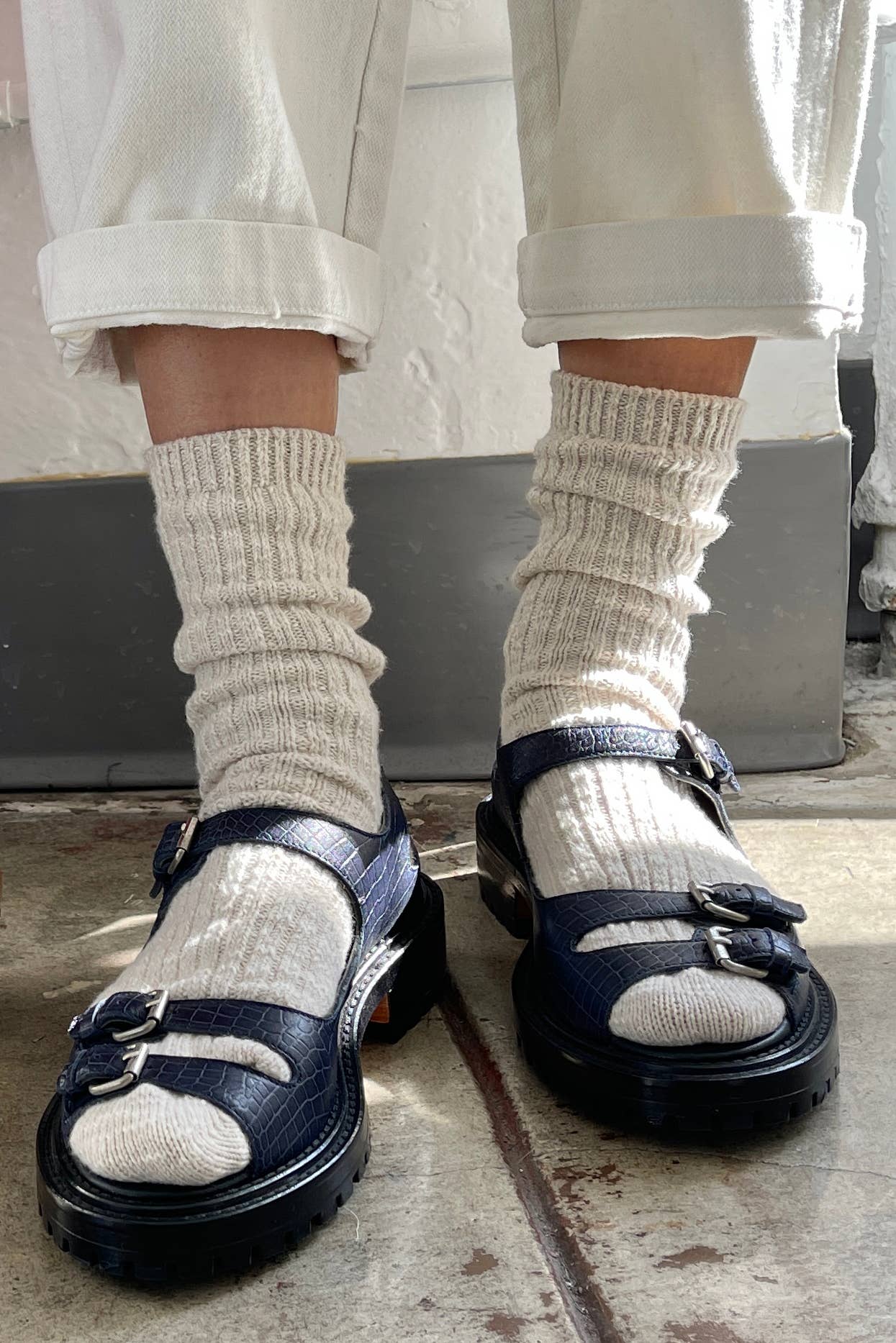 Person wearing navy sandals with beige Le Bon Shoppe  socks and rolled-up pants on a concrete floor.