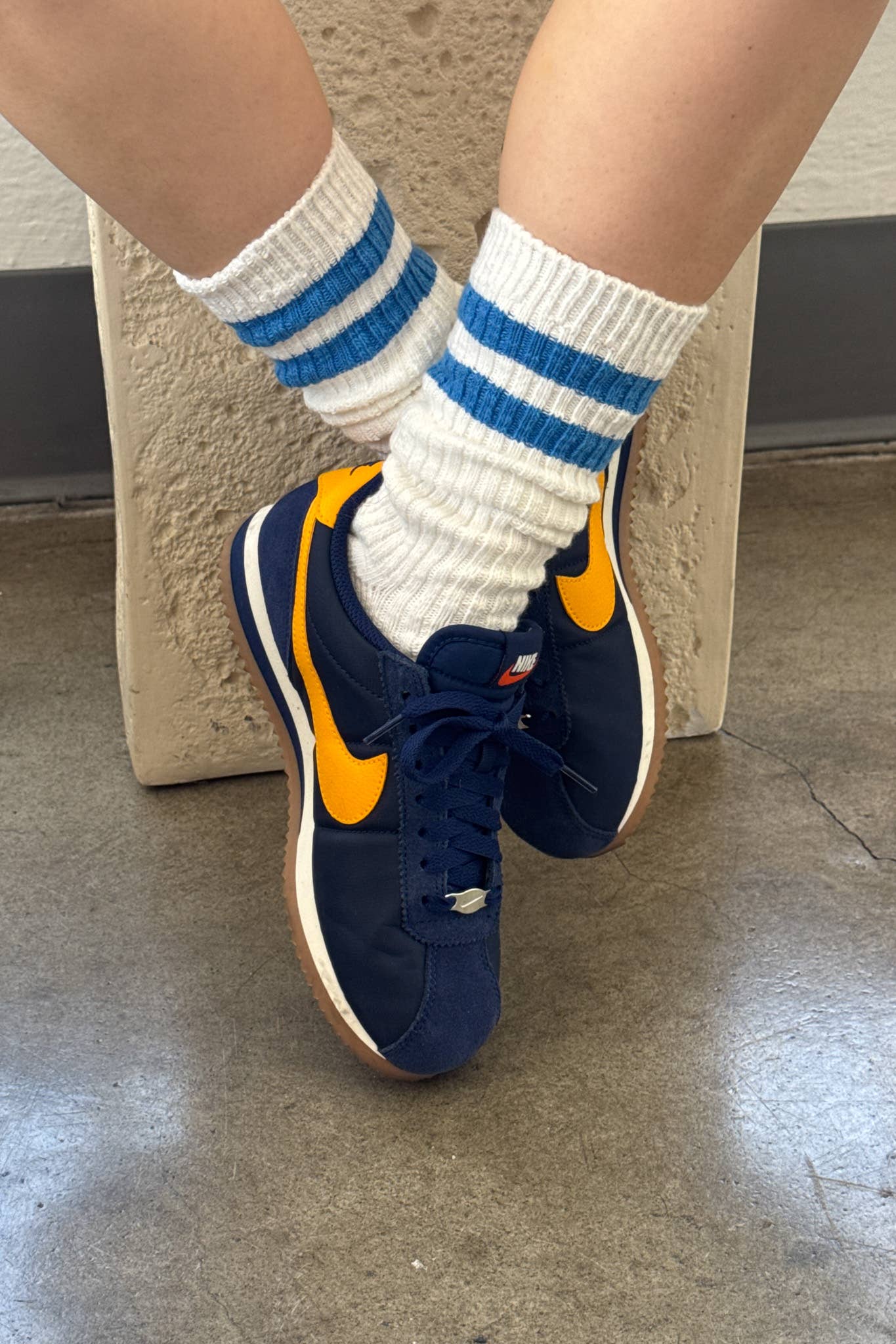 Blue sneakers with orange accents worn with Le Bon Shoppe white socks featuring blue stripes on a concrete floor.