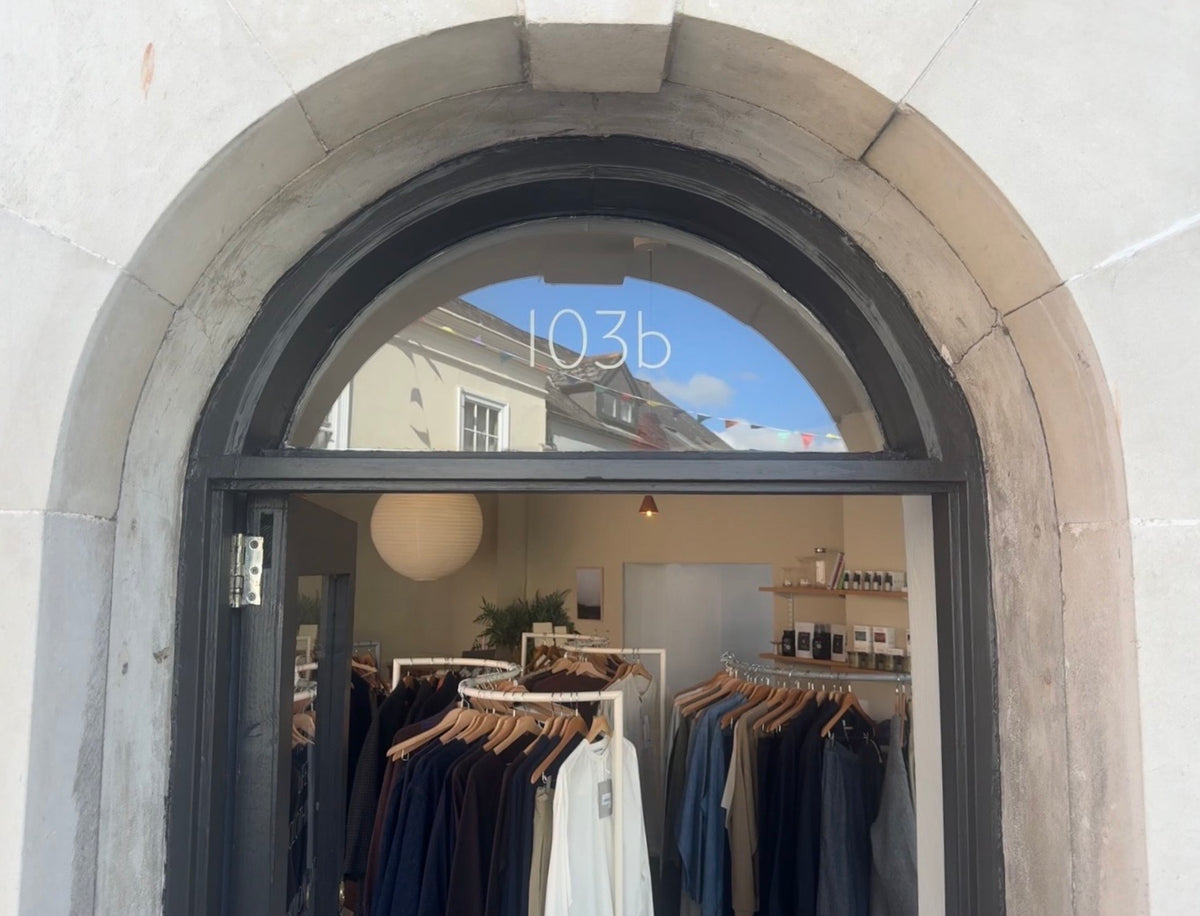 The storefront of a clothing boutique featuring a large arched window with the text '103b' printed on it. Inside, racks of clothing in navy, blue, and white are visible against a beige interior wall.