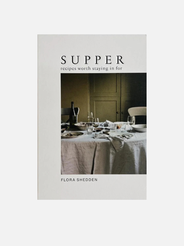A book cover for 'SUPPER: recipes worth staying in for' by Flora Shedden, featuring a minimalist off-white background with a central photograph of a dimly lit dining table set for a meal.
