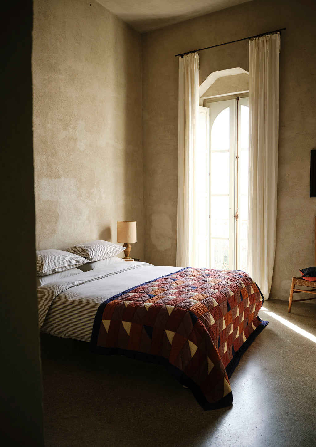 Bedroom with a bed covered by a patterned quilt, lamp, and large window with curtains.
