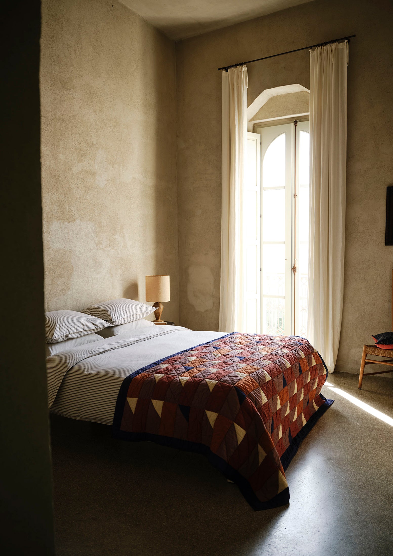 Bedroom with a bed covered by a patterned quilt, lamp, and large window with curtains.
