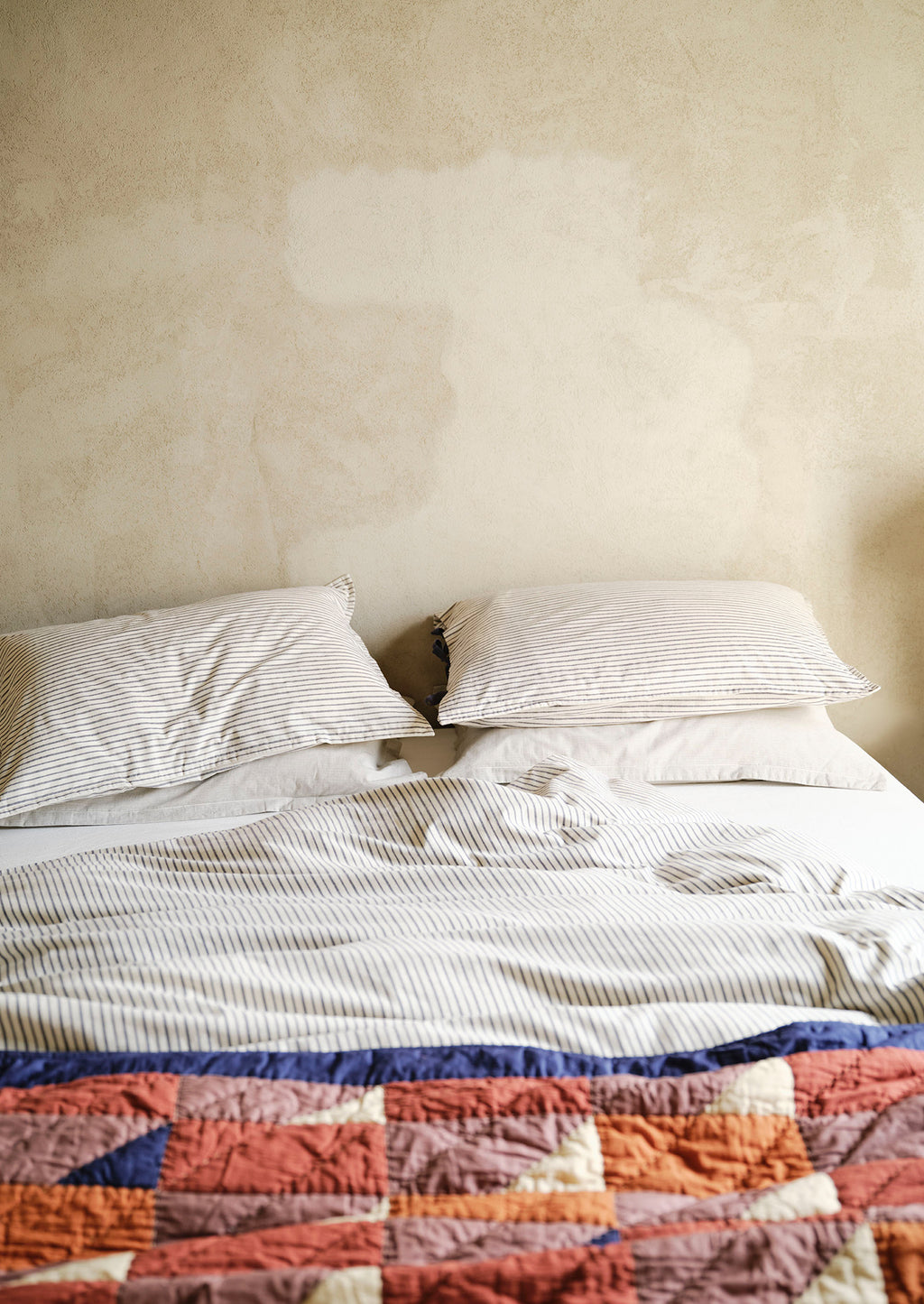 Bed with striped bedding and a colourful quilt against a plain wall.