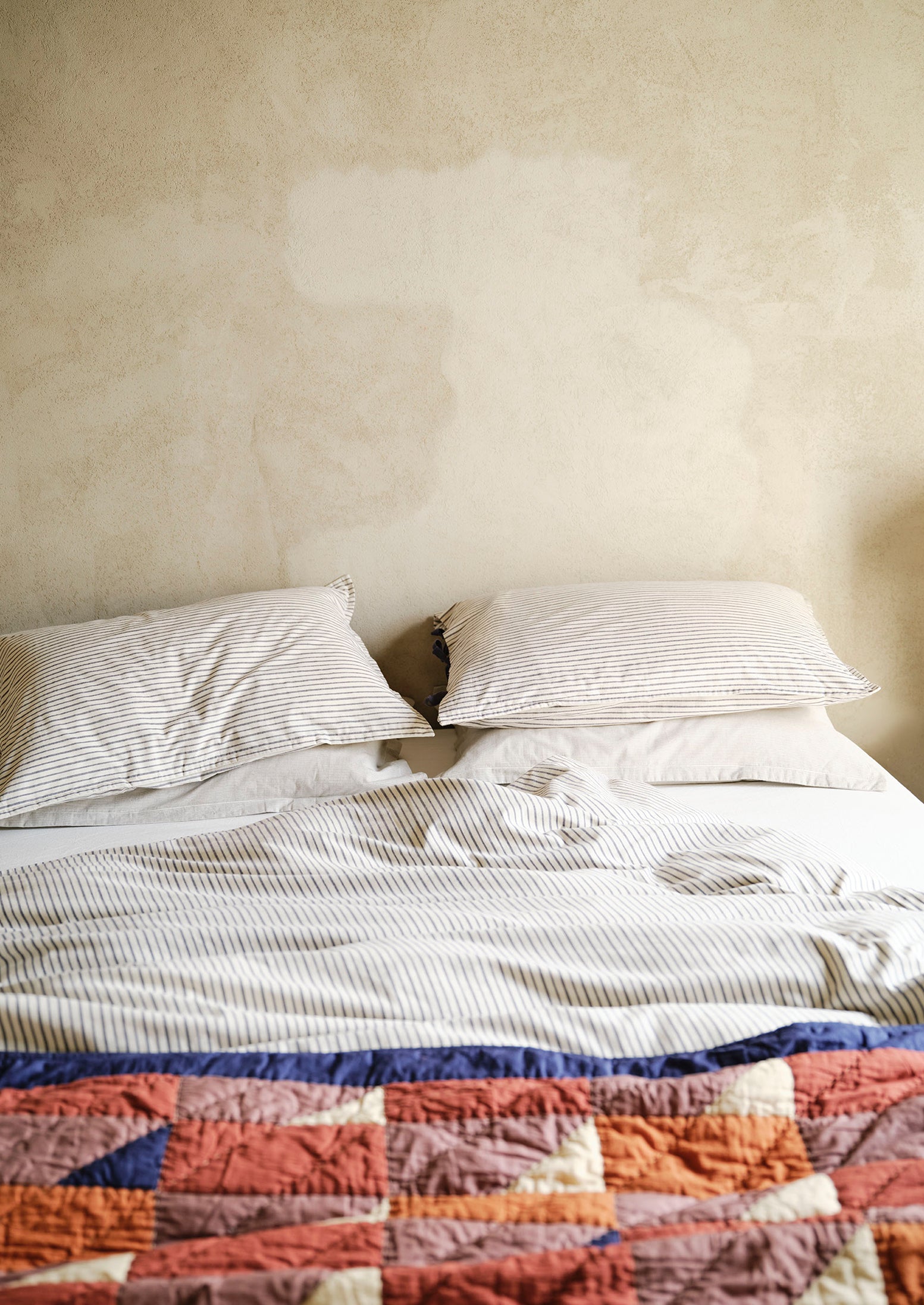 Bed with striped bedding and a colourful quilt against a plain wall.