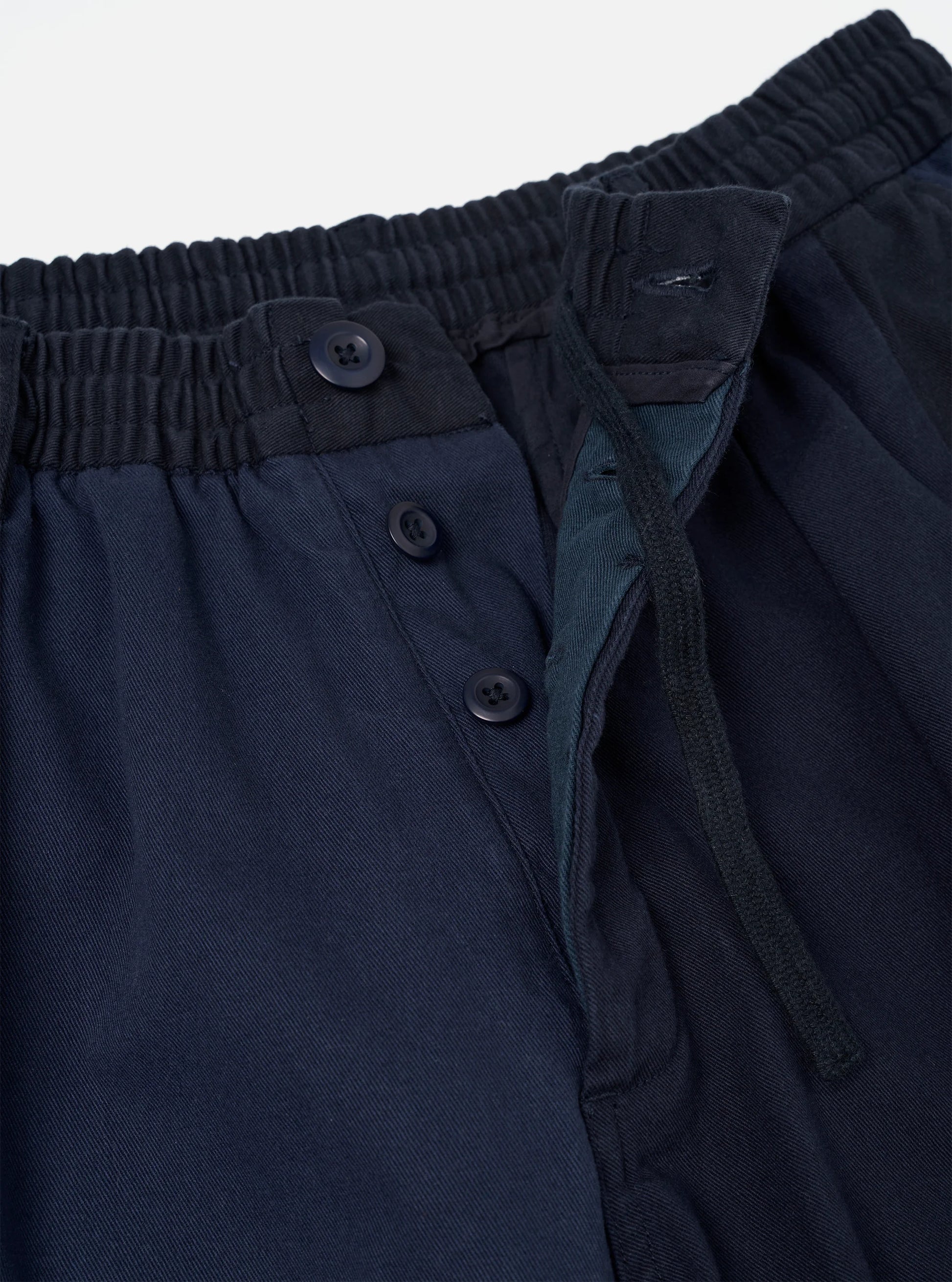 Universal Works Navy Twill patchwork Oxford pant close up fly detail against a light grey background. 