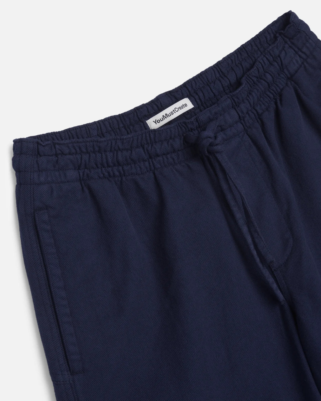 Close-up of dark navy blue drawstring trousers showing the elastic waistband and a white inner label that reads 'You Must Create'