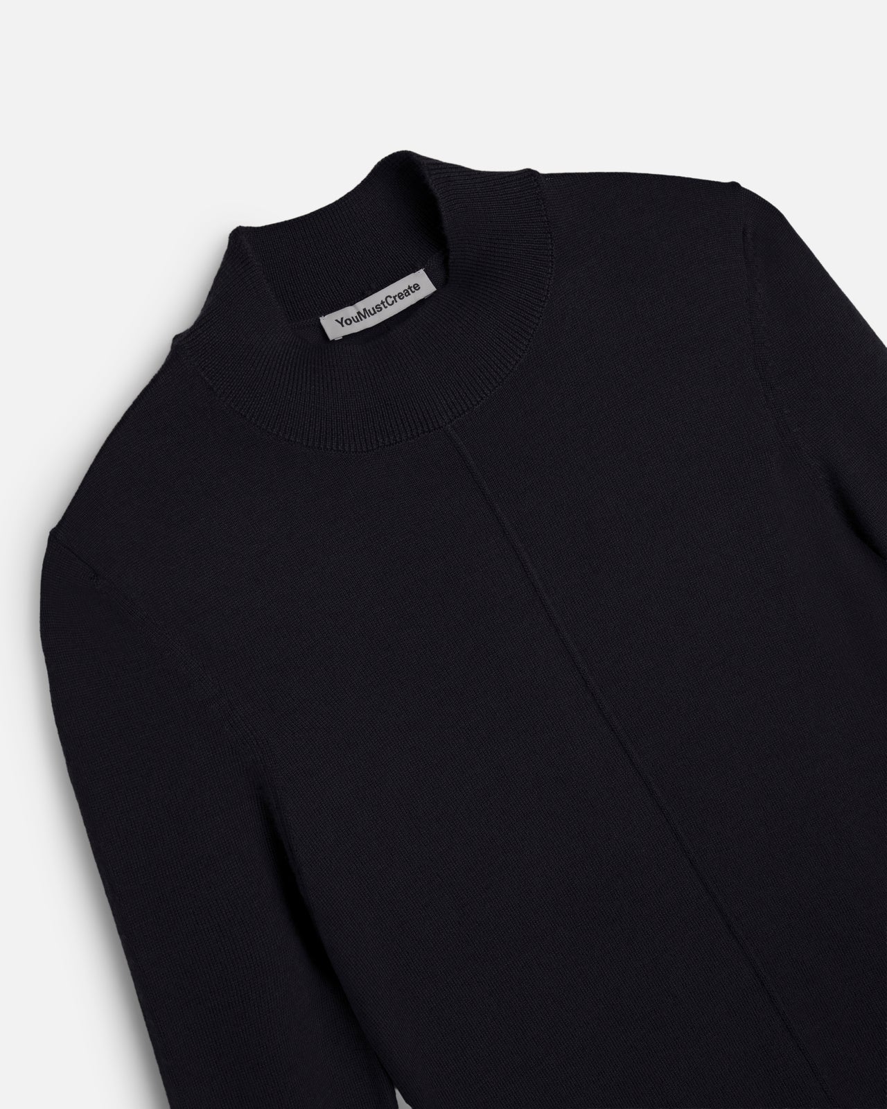 A close-up of a black knit mock neck YMC Youth Top with a visible 'You Must Create' label on the inside collar, shot against a light gray background.
