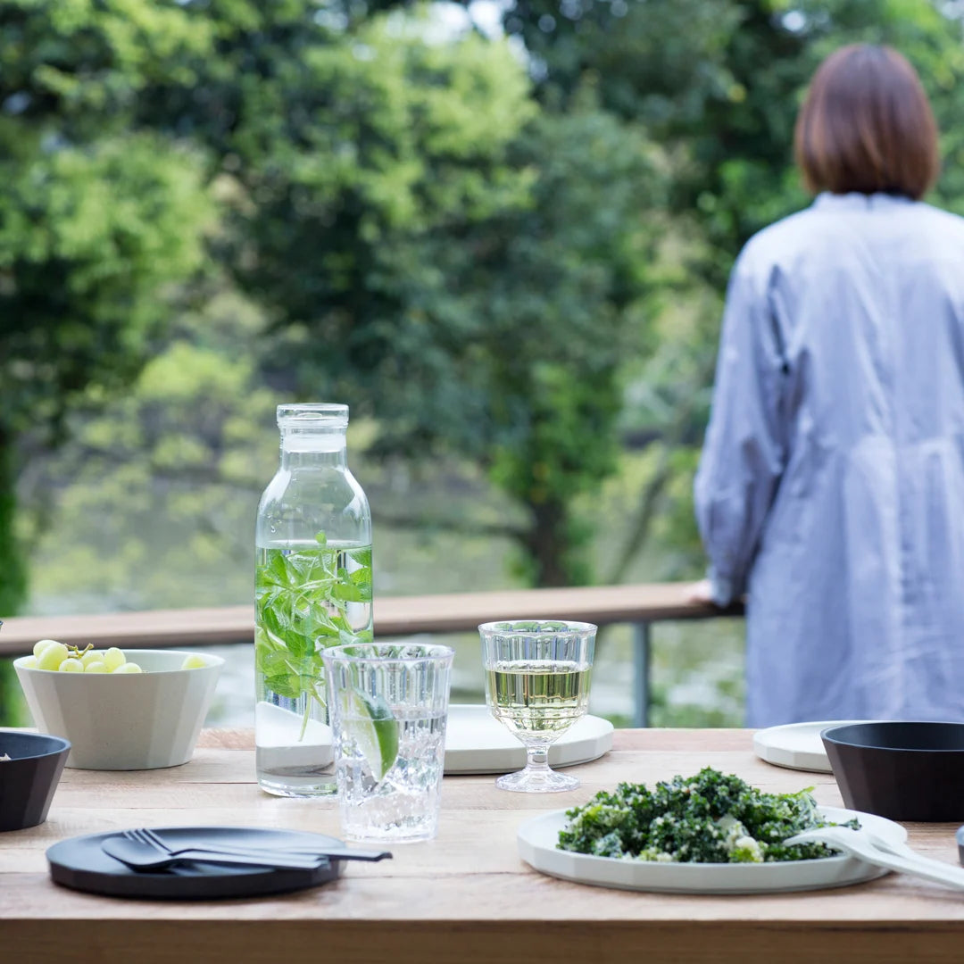 Person standing by a table with a meal and drinks, surrounded by greenery