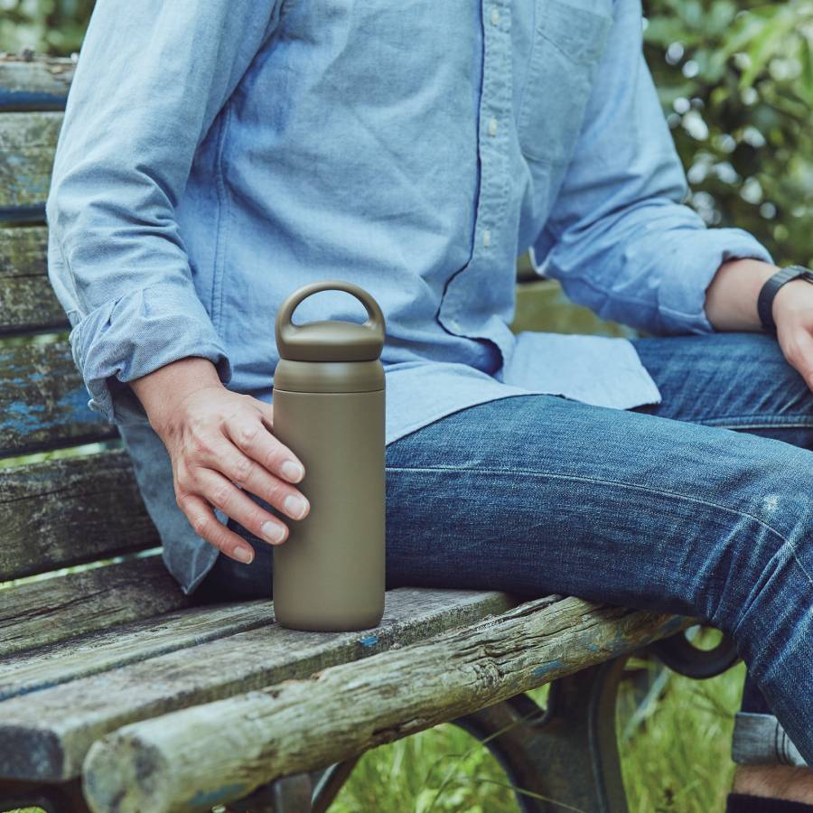 Person sitting on a wooden bench holding a brown insulated bottle outdoors.