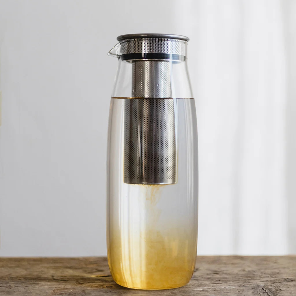 Clear carafe with gradient yellow color and metal lid on a wooden surface.